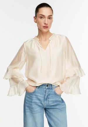 Femme aux cheveux lissés en arrière portant un chemisier crème à volants et un jean taille haute bleu clair, mains dans les poches, expression neutre.