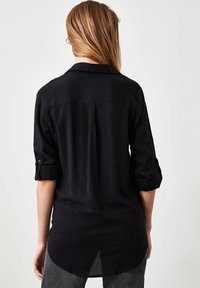 Camicia nera a maniche lunghe con polsini arrotolati, tessuto morbido, dettaglio piega posteriore e orlo curvo, vista posteriore.