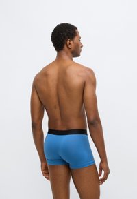 Boxer bleu avec une ceinture noire, design ajusté, texture lisse et détails de couture minimaux, porté par un modèle de dos.
