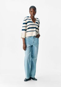 Gestreifter V-Ausschnitt-Pullover in Creme und Marineblau, mit Dreiviertelärmeln und Knopf-Details. Dazu hellblaue weite Jeans.