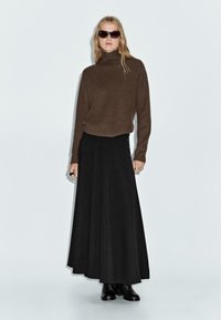 Frau mit langen blonden Haaren trägt einen braunen, gerippten Rollkragenpullover, einen schwarzen langen Rock, schwarze Stiefel, Sonnenbrillen und Ringe und steht vor einem schlichten Hintergrund.