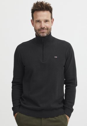Zwarte rits-zip sweater van gebreide stof, met een hoge kraag, geribde manchetten en een klein logo met rode, witte en blauwe accenten.
