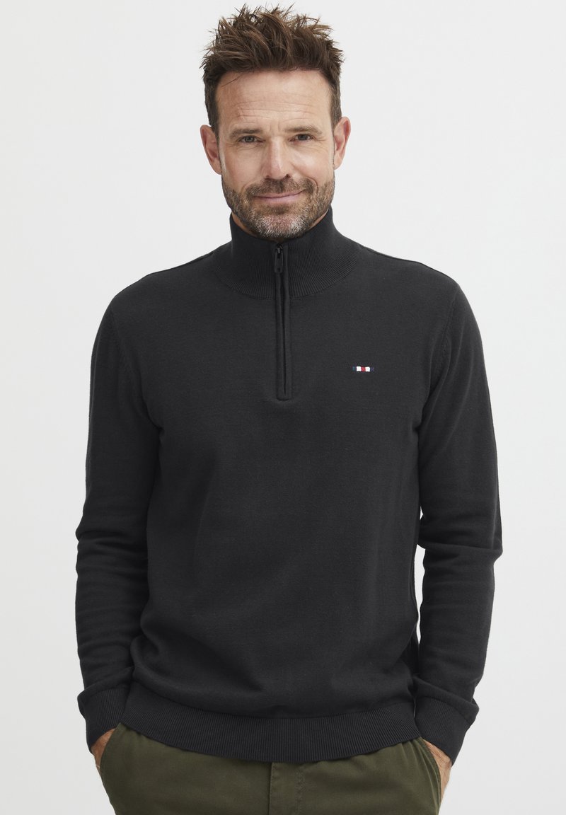 Sudadera negra con cremallera fabricada en tejido de punto, que presenta un cuello alto, puños acanalados y un pequeño logo con acentos en rojo, blanco y azul.