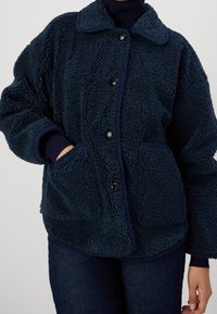 Granatowa polarowa kurtka o teksturowanej, puszystej powierzchni, z kołnierzem, przednimi kieszeniami i czarnymi guzikami. Luźny, oversize'owy krój.