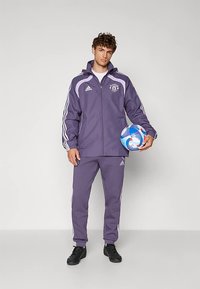 Paarse sporttrainingspak met witte strepen, bestaande uit een jas met rits en bijpassende broek. Model houdt een meerkleurige voetbal vast.