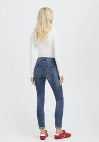 Sötétkék farmerból készült, magas derekú, karcsúsított nadrág, finoman kopott hatással és hátul zsebekkel. Piros cipővel és fehér hosszú ujjú felsővel párosítva.