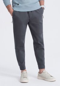 Pantalones de chándal grises con un ajuste entallado, que cuentan con bolsillos laterales, puños elásticos y detalles de paneles cosidos, combinados con zapatillas de color claro.