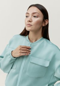 Jasnoniebieska kurtka bomberka wykonana z miękkiego materiału, z zamkiem błyskawicznym z przodu, elastycznymi mankietami i dwiema kieszeniami na piersiach z klapkami.