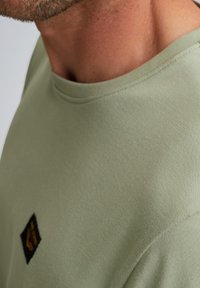 Krátky rukáv zelené tričko vyrobené z hladkej bavlny, s okrúhlym výstrihom a čiernou nášivkou loga na ľavej strane.