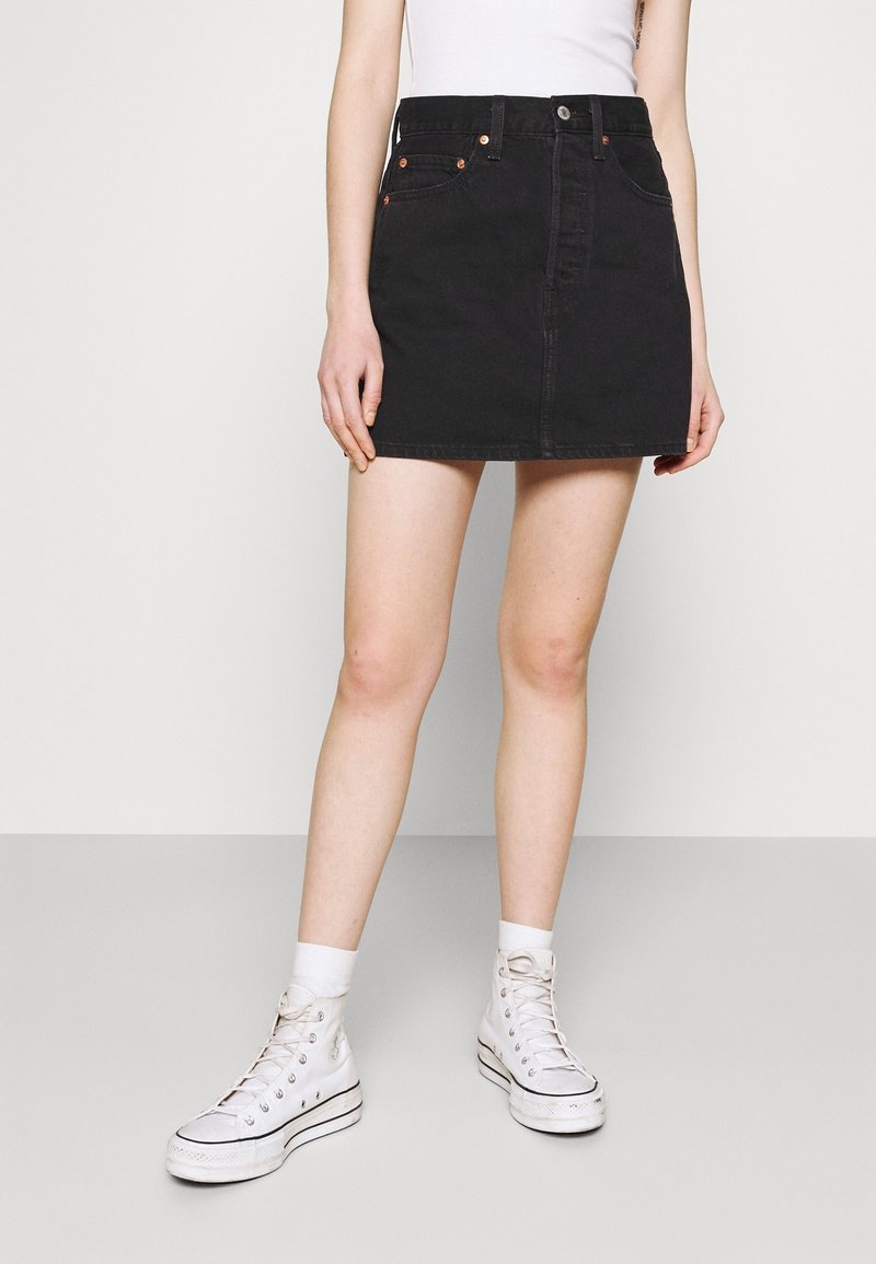 Jupe en denim noir à coupe droite, avec fermeture boutonnée sur le devant et poches. Portée avec des baskets blanches montantes et des chaussettes.