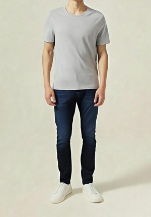 Homme portant un t-shirt uni gris clair à manches courtes, un jean bleu foncé et des chaussures blanches slip-on, debout devant un fond neutre.
