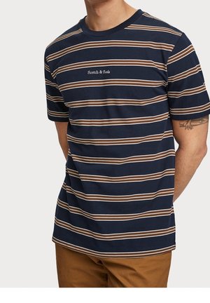 Mężczyzna w granatowej koszuli z krótkim rękawem z brązowymi i białymi poziomymi paskami oraz logo "Scotch & Soda" na klatce piersiowej, w połączeniu z brązowymi spodniami.