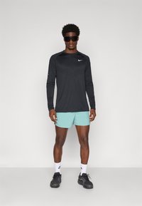 Zwarte langemouwen shirt met een gestructureerd ontwerp, lichtblauwe shorts, zwarte sneakers en donkere zonnebril, staand tegen een neutrale achtergrond.