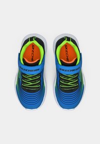 Kék sportcipők fekete kiegészítőkkel, textúrált felső résszel, zöld fűzőkkel és fehér talppal. A "SKECHERS" logó jól láthatóan elhelyezve.