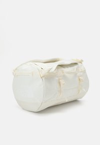 Torba za duffel u svijetloj krem boji, izrađena od izdržljivog materijala. Ima više remena, kopči i istaknuti logotip sa strane. Okrugli oblik.