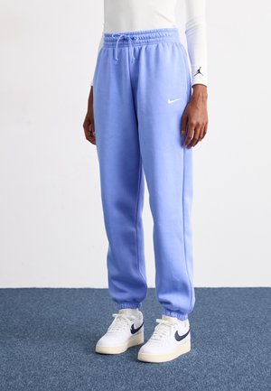 Persoon draagt lichtblauwe Nike joggingbroek met elastische boorden en trekkoord, wit langermig shirt en witte Nike sneakers met zwarte swoosh.