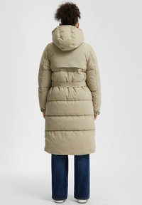 Dolga, podložena zunanja jakna v bež barvi z kapuco, pasom in vodoravnimi šivi. Kombinirana z širokimi modrimi kavbojkami in belimi čevlji.