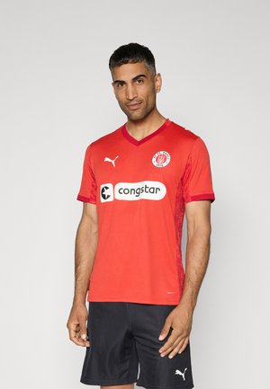 Maillot de football rouge à manches courtes avec col en V, panneaux latéraux texturés et logo blanc "congstar" et St. Pauli. Short de sport noir.