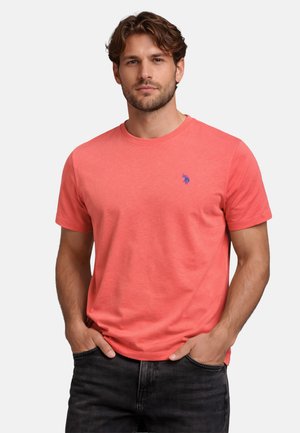 Man with light skin and brown hair wearing a coral short-sleeve T-shirt with a small blue logo and black jeans, hands in pockets.