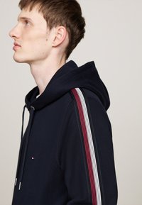 Sudadera azul marino con cordón, rayas laterales en rojo, blanco y negro, y un pequeño logo en el pecho. Textura suave y corte casual.