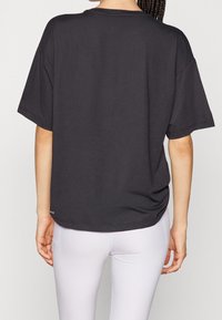 Černé oversized tričko s kulatým výstřihem, krátkými rukávy a hladkou texturou. Lem má jemný detail se logem blízko spodního okraje.