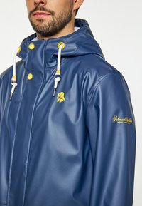 Mornarsko modra nepremočljiva jakna iz sijajnega materiala, z kapuco, rumenimi poudarki in logotipom na levem prsnem delu. Vključeni so beli žepi.
