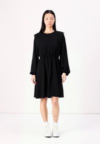 Black long-sleeve dress featuring lace detailing on shoulders, elastic waist, and a flared skirt. Paired with white high-top sneakers.