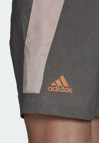 Harmaat urheilushortsit, joissa on kevyt, teksturoitu kangas, vaaleanpunainen sivupaneeli ja oranssi Adidas-logo helmassa.