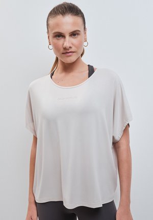 Giovane donna con capelli chiari raccolti, indossa una maglia bianca a maniche corte e larga sopra un reggiseno sportivo nero, in piedi davanti a uno sfondo grigio uniforme.