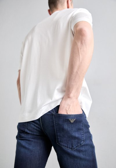 Weißes Kurzarm-T-Shirt, kombiniert mit dunkelblauen Jeans aus Denim, mit einem Metall-Logo-Akzent auf der Gesäßtasche und einem taillierten Design.