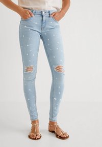 Jeans ajustados de color azul claro con un diseño salpicado de pintura blanca, detalles de rodillas desgastadas, cintura alta y estilo de cinco bolsillos.