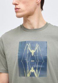 Νεαρός άνδρας που φοράει γκρι μπλουζάκι με κίτρινο αφηρημένο σχέδιο αράχνης και τη λέξη «BOSS» σε μπλε τετράγωνο φόντο.