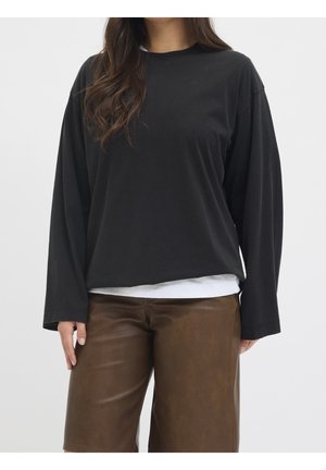 Femme portant une chemise noire ample à manches longues par-dessus un top blanc, associée à un short en cuir marron longueur genoux, debout devant un fond uni.