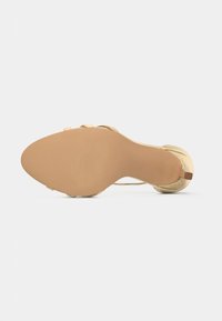 Pohja yksittäisestä metallinhohtoisesta kultaisesta sandaalista, jossa on matala korko ja ohut nilkkaremmiosa valkoista taustaa vasten.