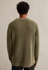 Olivově zelený žebrovaný pletený svetr s dlouhými rukávy, kulatým výstřihem a volným střihem. Obsahuje strukturovanou látku a mírně zakřivený lem.