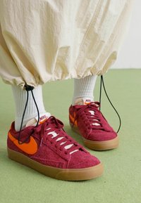 Zapatillas de ante burdeos con el swoosh naranja de Nike, cordones blancos y suelas de goma. Llevadas con pantalones crema y calcetines blancos, sobre un suelo verde.