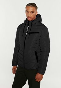 Veste noire rembourrée avec fermeture éclair, capuche et étiquette de marque. Comprend une poche poitrine zippée et des poches latérales, texture lisse et design léger.