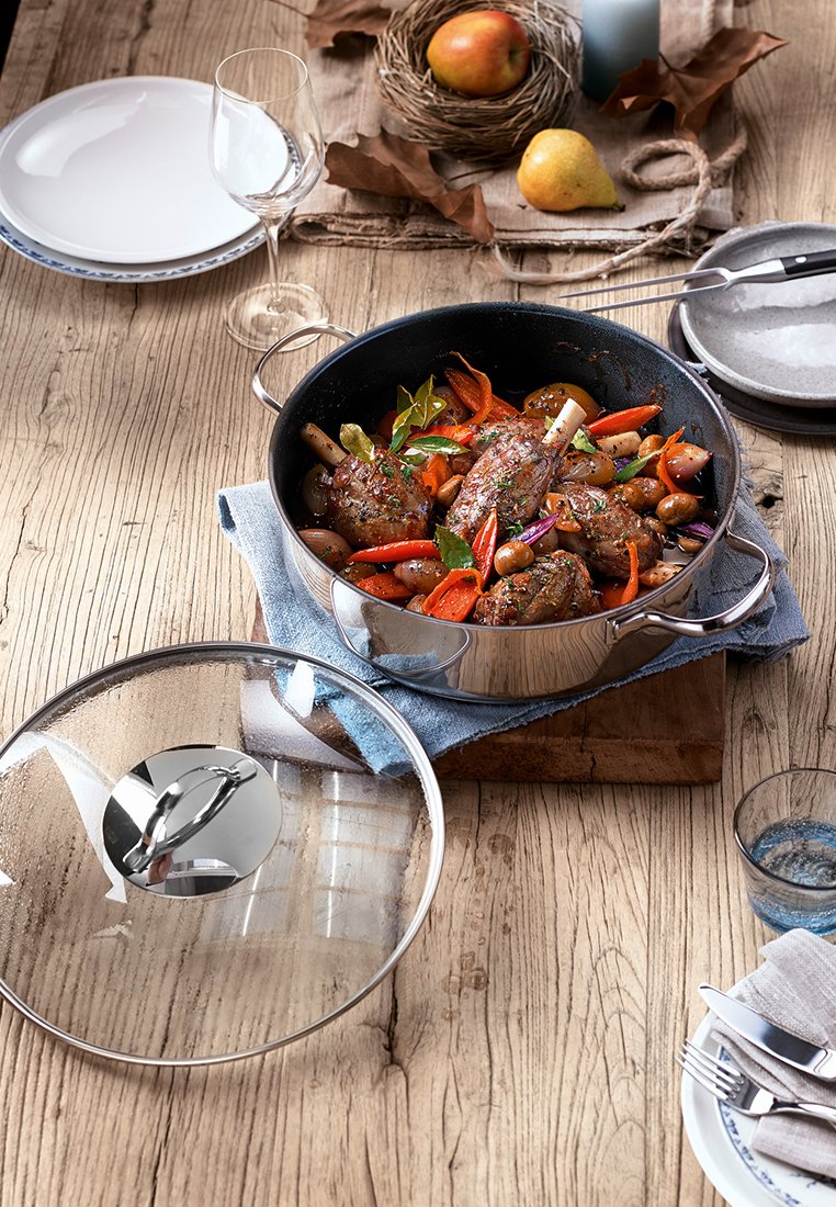 Gusseisen-Topf mit einem Glasdeckel, gefüllt mit buntem Gemüse und Fleisch. Holzoberfläche, blaue Stoffe, Keramikteller und Glaswaren im Hintergrund.