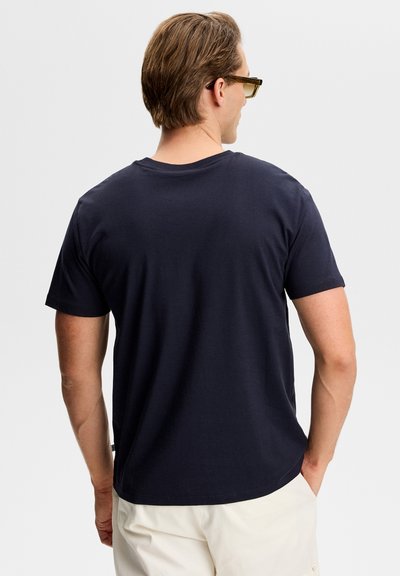 Άνδρας με ανοιχτό καστανά μαλλιά, φοράει σκούρο μπλε μπλουζάκι και κρεμ παντελόνι, γυρισμένος με την πλάτη, με τα χέρια στις τσέπες μπροστά σε απλό ανοιχτό φόντο.