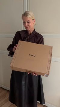 Rjava kartonska škatla z napisom "MNG" v črni barvi. Škatlo drži oseba v črni, povešeni usnjeni krilu in bluzici z nabranimi detajli.