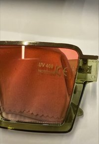 Sonnenbrille mit goldenem Rahmen und orange getönten Gläsern, auf denen der Text "UV 400 Schutz" zu sehen ist, sowie einem orangefarbenen Reinigungstuch hinter den Gläsern.