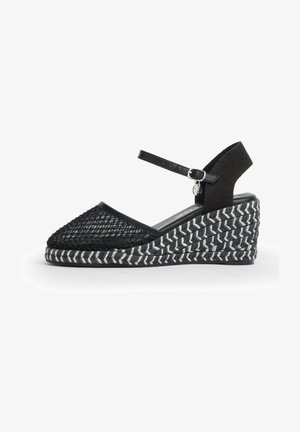 Zapato de cuña negro con puntera cerrada tejida, correa en el tobillo con hebilla y suela texturizada con patrón espiga en blanco y negro.