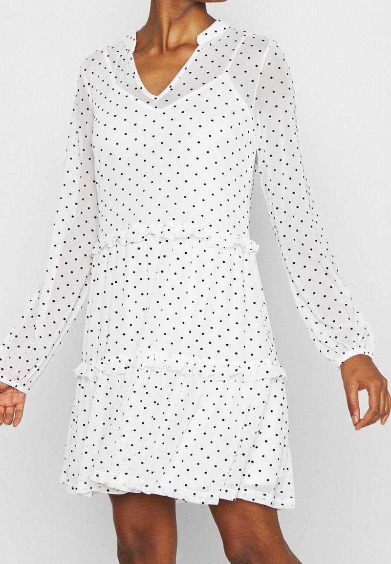 Robe blanche à manches longues avec des pois noirs, des volants à la taille et à la jupe, col en V, portée par une personne à la peau brune.