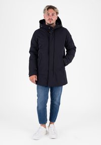 Marine parka met een capuchon, ritssluiting aan de voorkant, twee voorzakken en een iets langere achterkant. Gedragen over lichtblauwe jeans en witte sneakers.