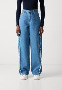 Weite blaue Denim-Jeans mit ausgefransten Seitenähten, hoher Taille und Gürtelschlaufen. Kombiniert mit weißen Slipschuhen.