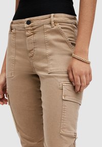 Beiga cargobyxor i bomullsmix med sidofickor, smal midja och synliga sömdetaljer. Händer som greppar midjebandet.