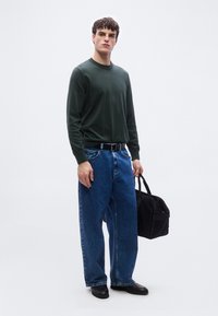 Νεαρός άνδρας που φοράει σκούρο πράσινο πουλόβερ, φαρδιά μπλε τζιν, μαύρη ζώνη και παπούτσια, κρατώντας μια μαύρη σακούλα duffel μπροστά σε ένα απλό φόντο.