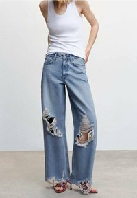 Jean large déchiré en denim bleu clair, avec de grandes déchirures aux genoux et des bords effilochés, associé à un débardeur blanc.
