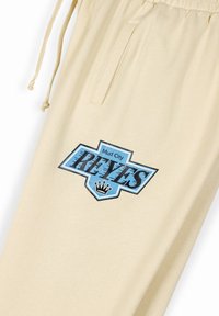 Beige Sweatpants mit einem blauen grafischen Logo, das "Mud City REYES" und eine Krone auf dem linken Oberschenkel zeigt; verfügt über einen Kordelzugbund und eine Seitenstasche.