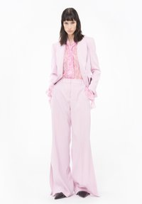 Completo su misura rosa chiaro con pantaloni a gamba larga e blusa trasparente rosa a balze. Scarpe nere con tacco alto completano l'outfit.
